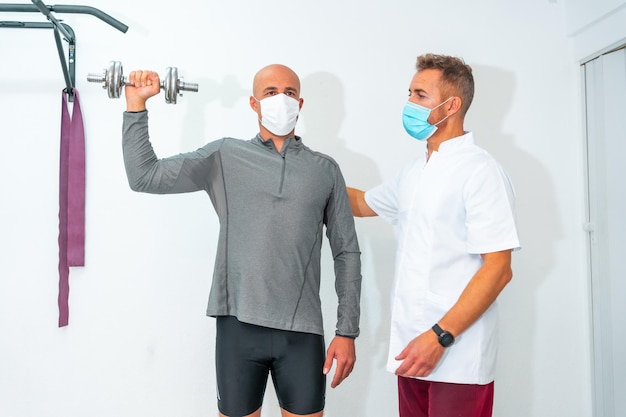 Fizjoterapeuta z maską na twarz i pacjent ćwiczący z hantlami Fizjoterapia ze środkami ochronnymi na wypadek pandemii koronawirusa COVID19 Osteopatia sportowy quiromassage