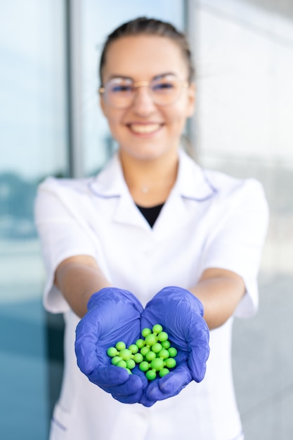 Fizyka, nauka, biologia i medycyna koncepcja - uśmiechnięta orientalna fizyczka w białym fartuchu, goglach i niebieskich gumowych rękawiczkach trzymająca zielone atomy na ulicy