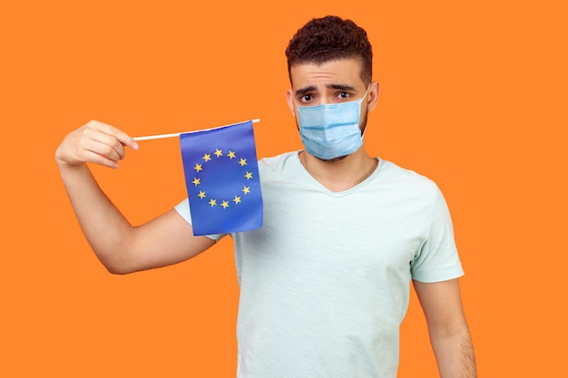 Flaga Unii Europejskiej Portret zdenerwowanego mężczyzny z maską medyczną w białej koszulce trzymającej flagę UE i patrzącego na kamerę z rozczarowanym smutnym wyrazem studio strzał na białym tle na pomarańczowym tle