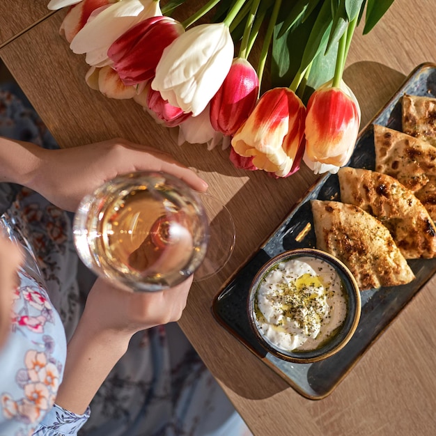 Flatlay kobiecej ręki trzymającej kieliszek wina i bukiet kwiatów na tle stołu widok z góry