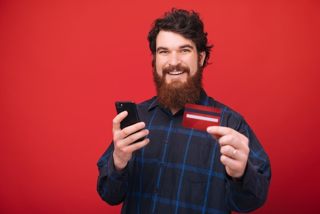 Fotografia pokazuje jego nową kredytową kartę przystojny mężczyzna podczas gdy używać wiszącą ozdobę nad czerwieni ścianą