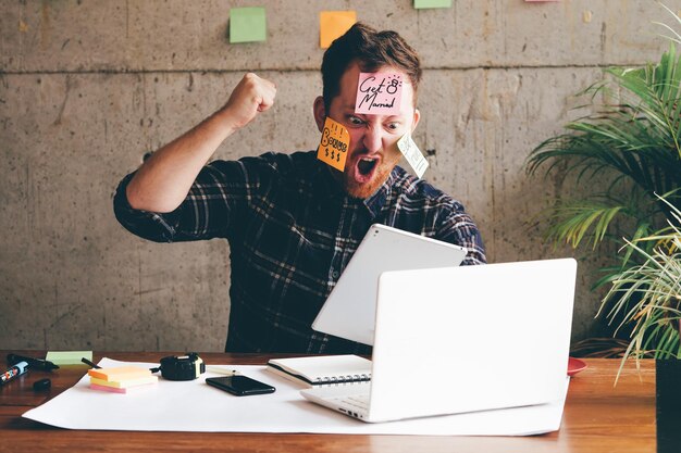 Zdjęcie frustrowany biznesmen z klejowymi notatkami na twarzy używający laptopa w biurze