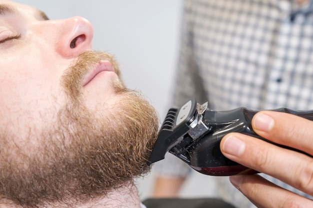 fryzjer goli brodę swojego klienta za pomocą trymera elektrycznego.