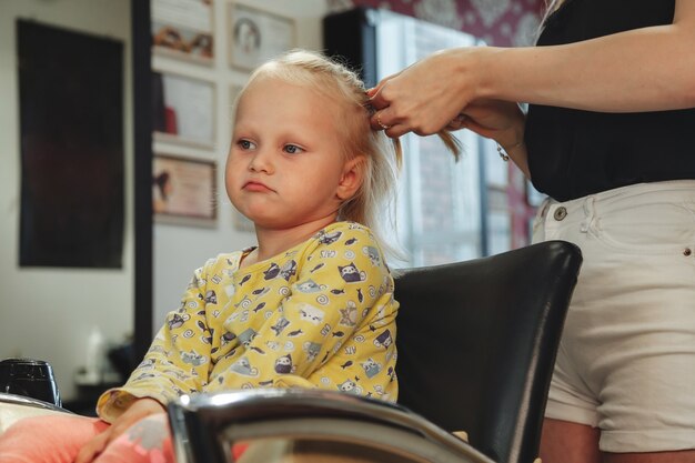 Fryzjer kobieta zrobić modną ładną fryzurę dla cute blond dziewczynka dziecko w nowoczesnym fryzjera, salon fryzjerski. Fryzjer sprawia, że fryzura dla małego dziecka w salonie fryzjerskim. Koncepcja fryzury i urody