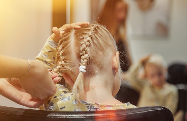 Fryzjer kobieta zrobić modną ładną fryzurę dla cute blond dziewczynka dziecko w nowoczesnym fryzjera, salon fryzjerski. Fryzjer sprawia, że fryzura dla małego dziecka w salonie fryzjerskim. Koncepcja fryzury i urody