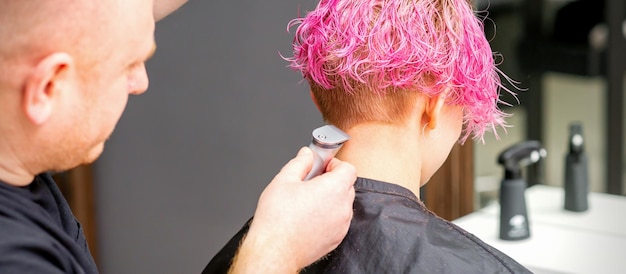 Fryzjer męski goli szyję młodej kobiety rasy kaukaskiej z krótką różową fryzurą golarką elektryczną w salonie fryzjerskim z bliska