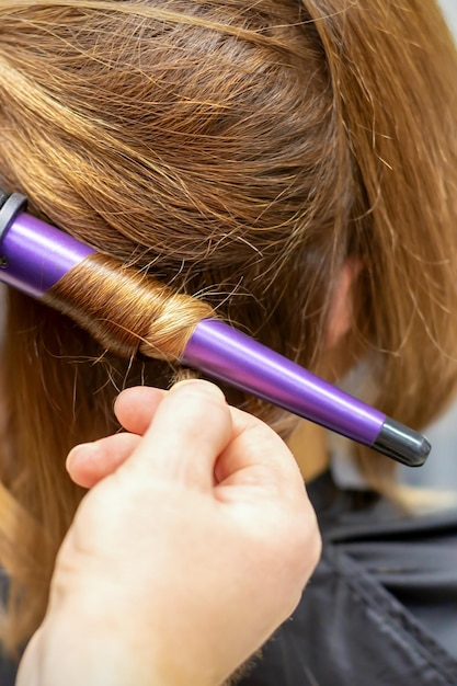 Fryzjer robi loki fryzurę długich brązowych włosów za pomocą lokówki w salonie fryzjerskim z bliska