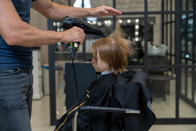 Fryzjer-stylista po strzyżeniu suszy włosy chłopca suszarką do włosów