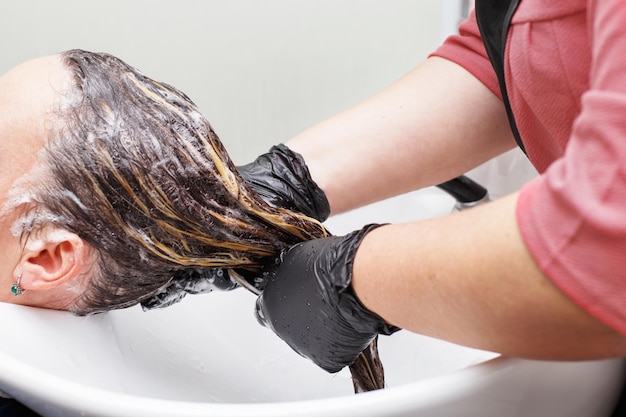 Fryzjer w czarnych rękawiczkach myjący włosy brunetki w salonie kosmetycznym