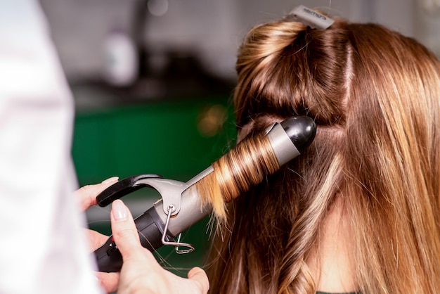 Fryzjerka kręci włosy brązowowłosej młodej kaukaskiej kobiecie w salonie kosmetycznym