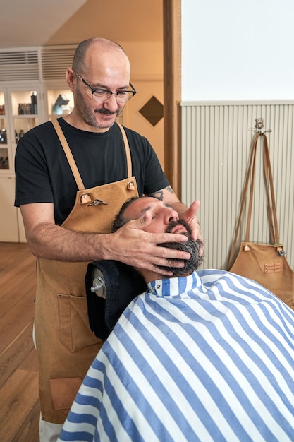 Fryzura dorosłego mężczyzny w odzieży roboczej stojąca w pobliżu klienta i nakładająca żel podczas zabiegu leczenia brody w nowoczesnym zakładzie fryzjerskim