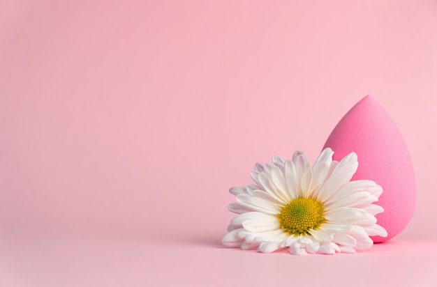Zdjęcie gąbka do aplikacji makijażu w formie kropli lub jajka oraz białej chryzantemy na delikatnym różowym tle.