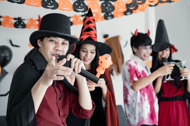 Gang młodych Azjatów w stroju czarownicy, czarodziej z imprezą Halloween za zaśpiewanie piosenki i drinka, deser w pokoju. Grupa nastolatków Thai z świętować Halloween. Koncepcja party Halloween w domu.