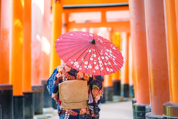 Gejsza ubrana w japońskie kimono wśród czerwonej, drewnianej bramy Tori w świątyni Fushimi Inari w Kioto