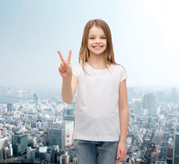 gest i koncepcja szczęśliwych ludzi - uśmiechnięta dziewczynka w białej pustej koszulce pokazująca gest pokoju palcami