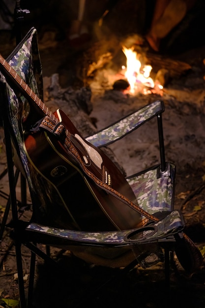 Gitara akustyczna w fotelu kempingowym z płomieniem ognia na tle. letni kemping. relaks na łonie natury. Obraz pionowy. wysoki poziom
