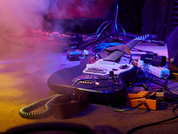 Gitara i sprzęt gitarowy leżą na scenie we mgle i dymią w fioletowym, niebieskim i pomarańczowym oświetleniu.