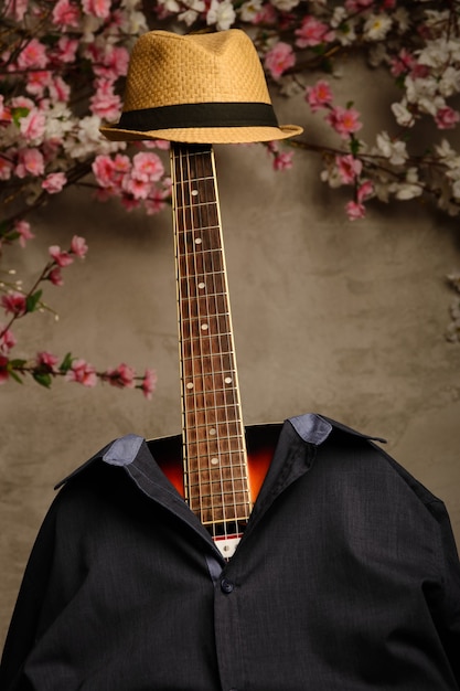 Gitara ubrana jest w męską niebieską koszulę i kapelusz z podstrunnicą przy ścianie kwiatów