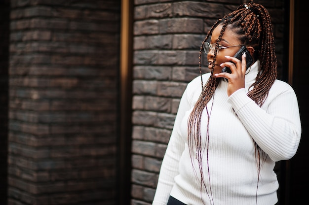 Glamorous African American kobieta w biały sweter z golfem pozuje na ulicy z telefonu komórkowego.
