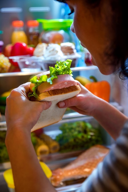 Głodna kobieta trzymająca w rękach kanapkę i stojąca obok otwartej lodówki. Niezdrowe jedzenie.