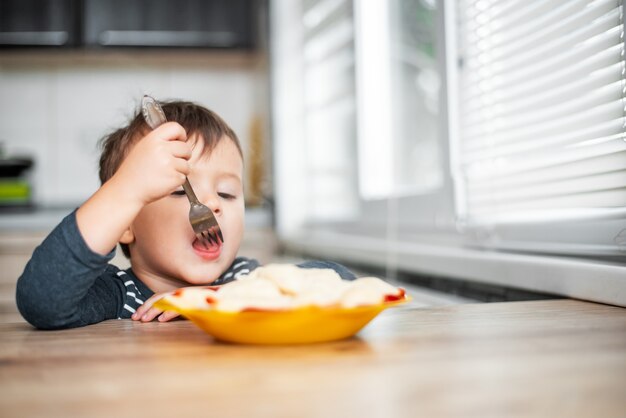 Głodne dziecko je pierogi w kuchni, siedząc przy stole w szarej kurtce