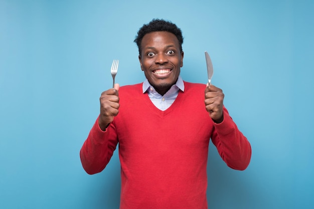 Głodny afrykański młody człowiek trzyma widelec i nóż pod ręką gotowy do jedzenia