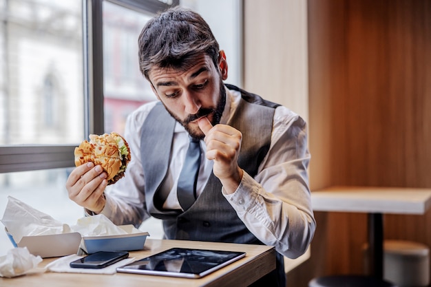 Głodny mężczyzna w garniturze siedzi w restauracji typu fast food w przerwie na lunch, je burgera z serem i czyta wiadomości na tablecie.