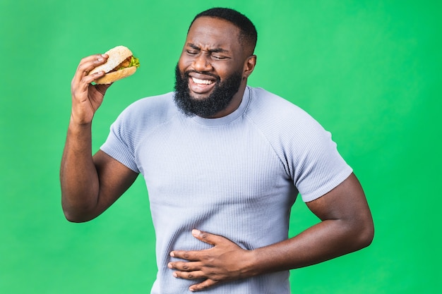 Głodny młody african american Murzyn jedzenie hamburgerów na białym tle nad zielonym tłem. Ból brzucha.