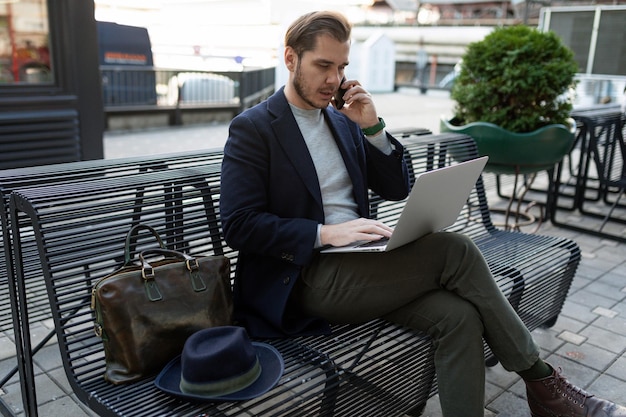 Głowa mężczyzny w średnim wieku pracuje na laptopie i rozmawia przez telefon komórkowy, siedząc na ławce