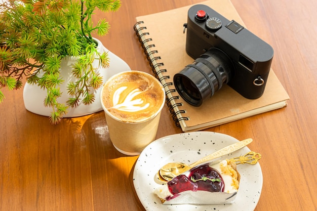 Gorąca kawa latte z pianką mleczną latte art w filiżance wykonanej z papieru i aparatu w notatniku Ciasto jagodowe na drewnianym biurku na widoku z góry jako śniadanie w kawiarni w kawiarnipodczas koncepcji pracy biznesowej