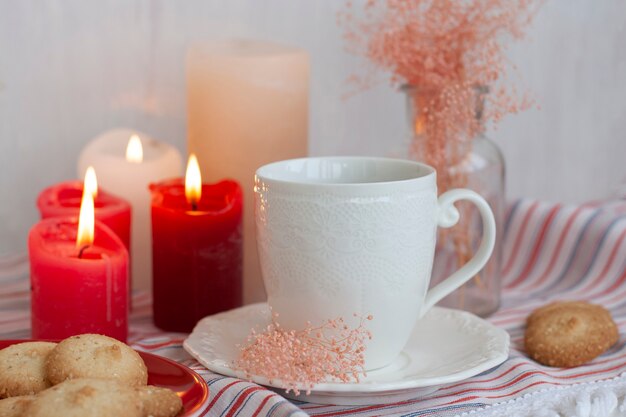 gorący kubek czarnej herbaty na obrusie w paski, świece woskowe, szklany wazon z dekoracyjnymi ziołami