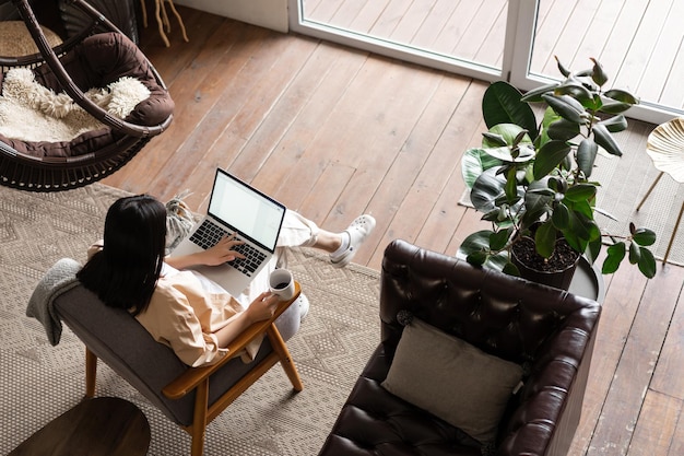 Górny kąt ujęcia zajętej azjatyckiej kobiety pracującej w domu na laptopie siedzącej w salonie i...