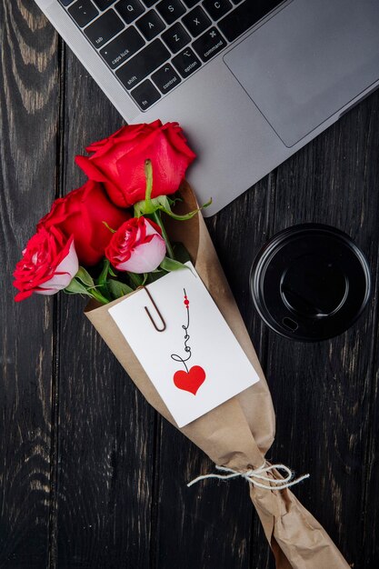 górny widok bukiet czerwonych róż w papierze rzemieślniczym z załączoną pocztówką leżący w pobliżu laptopa z papierowym kubkiem kawy na ciemnym drewnianym tle