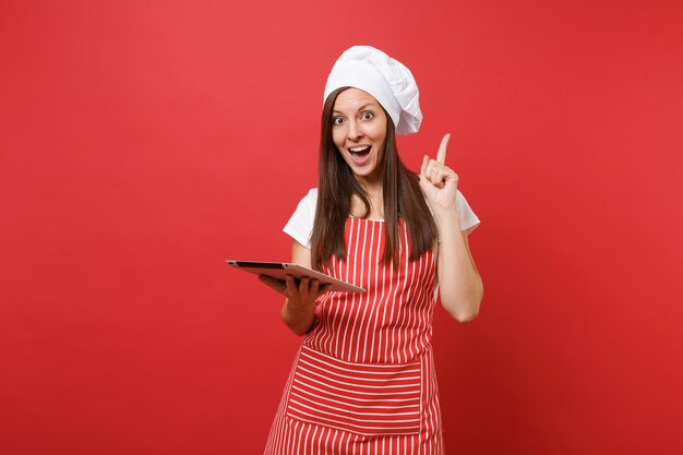 Gospodyni domowa żeński kucharz lub piekarz w pasiasty fartuch, biała koszulka, kapelusz kucharzy toczek na białym tle na tle czerwonej ściany. Zabawna gospodyni kobieta szuka przepisu na komputerze typu tablet. Makieta koncepcja przestrzeni kopii.