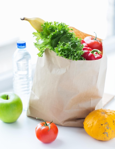 Zdjęcie gotowanie, dieta, wegetariańskie jedzenie i koncepcja zdrowego odżywiania - zbliżenie papierowej torby ze świeżymi dojrzałymi soczystymi owocami i warzywami oraz butelką wody na kuchennym stole w domu