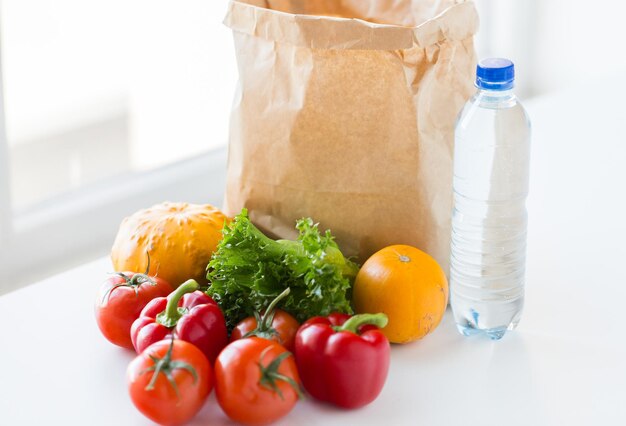 Zdjęcie gotowanie, dieta, wegetariańskie jedzenie i koncepcja zdrowego odżywiania - zbliżenie papierowej torby ze świeżymi dojrzałymi soczystymi warzywami i butelką wody na kuchennym stole w domu