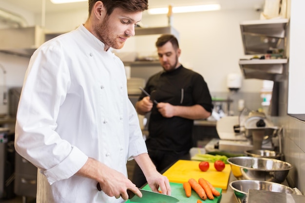 gotowanie jedzenia, zawód i koncepcja ludzi - szczęśliwy szef kuchni z nożem siekającym marchewkę na desce do krojenia i kucharzem do ostrzenia noża w kuchni restauracji