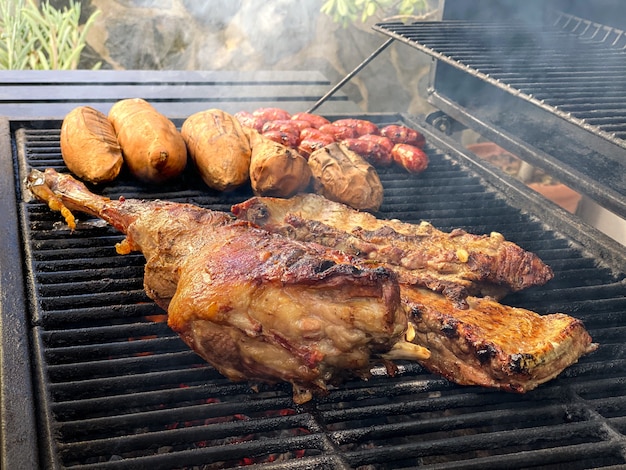 Gotowanie pysznego udźca jagnięcego na metalowej tacy grillowanej. Na grillu BBQ ugotuj kiełbasę z mięsa chorizo i batatów. Jedzenie gotowane z grillem.