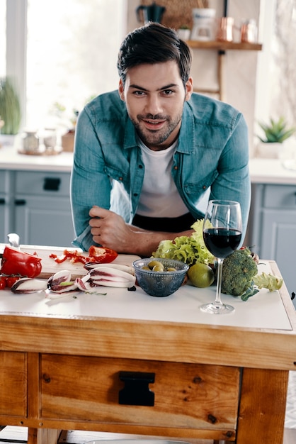 Gotowanie zdrowego obiadu. Przystojny młody mężczyzna w stroju casual patrząc na kamerę i uśmiechając się stojąc w kuchni w domu