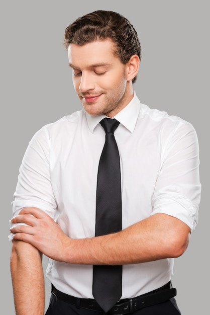Zdjęcie gotowy do pracy. pewny siebie młody człowiek w koszuli i krawacie, dopasowując rękawy, stojąc na szarym tle