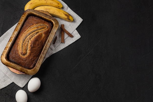 Gotowy świeży chleb bananowy w prostokątnym naczyniu do pieczenia ze składnikami na ciemnym tle