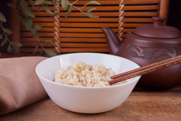 Gotujący brown ryż w białym pucharze z chopsticks na drewnianym tle.