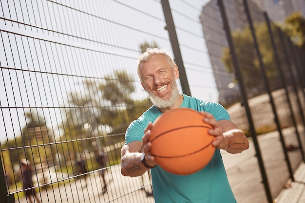 Zdjęcie gra w koszykówkę wesoły mężczyzna w średnim wieku w stroju sportowym, trzymający piłkę do koszykówki i uśmiechający się do