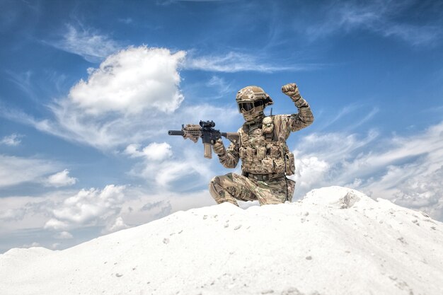 Gracz airsoftowych gier wojskowych w mundurze kamuflażowym i uzbrojonej w hełm repliki karabinu szturmowego, siedzący na kolanie na szczycie wydmy z niebem w tle i pokazujący zamrożony sygnał ręką swoim kolegom z drużyny