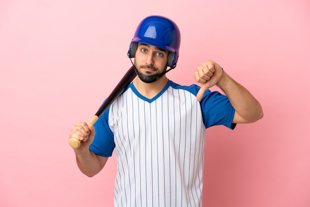 Gracz baseballa z hełmem i kijem na białym tle na różowym tle pokazując kciuk w dół z negatywnym wyrazem twarzy