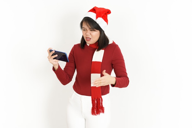 Grając w grę mobilną z piękną Azjatką ubraną w czerwony golf i czapkę Świętego Mikołaja na białym tle