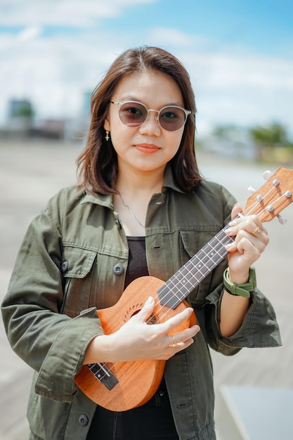 Grająca Na Ukulele Młoda Piękna Azjatka Ubrana W Kurtkę I Czarne Dżinsy Pozuje Na Zewnątrz