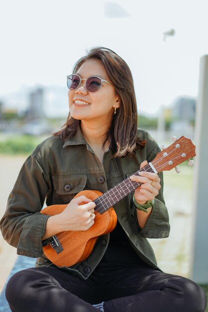 Grająca na ukulele młoda piękna Azjatka ubrana w kurtkę i czarne dżinsy pozuje na zewnątrz