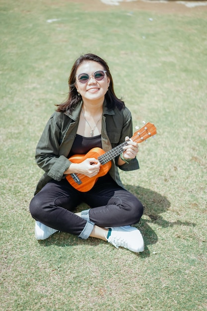 Grająca na ukulele młoda piękna Azjatka ubrana w kurtkę i czarne dżinsy pozuje na zewnątrz
