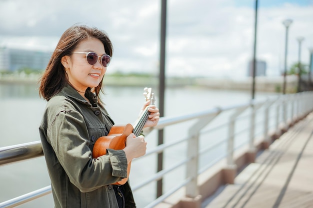 Grająca na ukulele młoda piękna Azjatka ubrana w kurtkę i czarne dżinsy pozuje na zewnątrz